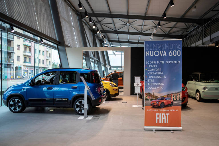 A imagem mostra uma exposição de carros da Fiat， com destaque para um carro azul à esquerda. Ao fundo， há outros carros em cores variadas， como laranja e verde. Um banner em destaque à direita apresenta a mensagem 039;BENVENUTA NUOVA 600039; e lista características como espaço， conforto， versatilidade e funcionalidade. O ambiente é moderno， com grandes janelas e iluminação adequada.