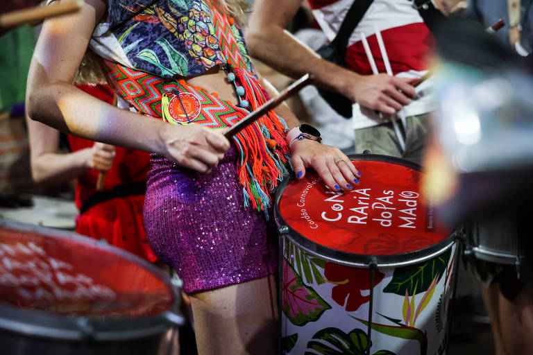 A imagem mostra uma pessoa tocando um tambor em um ambiente festivo. A pessoa está vestida com uma blusa colorida e uma saia brilhante, segurando uma baqueta. O tambor tem uma tampa vermelha com o texto 'Goiânia Raízes do Pandeiro'. Ao fundo, outras pessoas também estão visíveis, algumas vestindo roupas de festa.