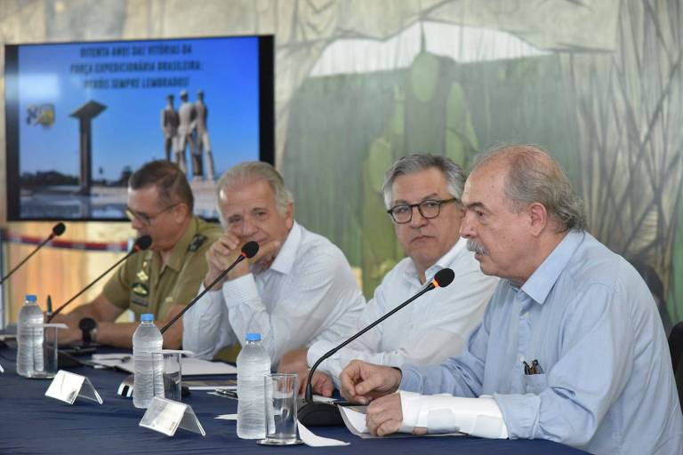 A imagem mostra uma mesa de reunião com quatro homens sentados. O primeiro à esquerda é um oficial militar， vestido com uniforme. Os outros três homens estão em trajes civis， com camisas de manga longa. Eles estão discutindo， com um microfone à frente de um deles. Ao fundo， há uma tela que exibe uma apresentação com imagens e texto relacionado ao 039;Prefeito Expedicionário Brasileiro039;.