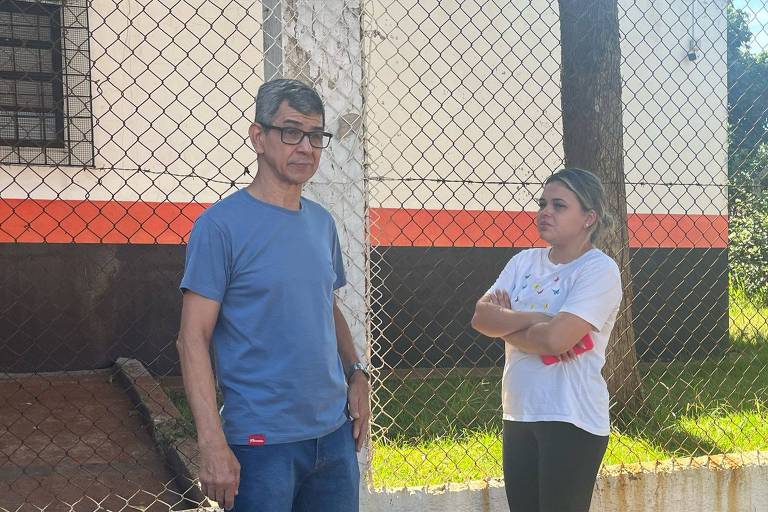 A imagem mostra um homem e uma mulher em pé， conversando em um ambiente externo. O homem， de cabelo grisalho e óculos， usa uma camiseta azul e está com as mãos nos bolsos. A mulher， com cabelo preso e uma camiseta branca， está com os braços cruzados， olhando para o homem. Ao fundo， há uma cerca e uma parede com pintura laranja na parte inferior.