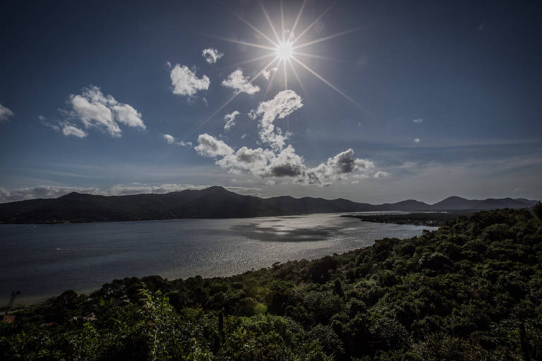 A imagem mostra uma vista panorâmica de um lago cercado por montanhas. O sol brilha intensamente no céu， criando reflexos na água. Nuvens brancas estão dispersas no céu azul， e a vegetação verde se estende na parte inferior da imagem.