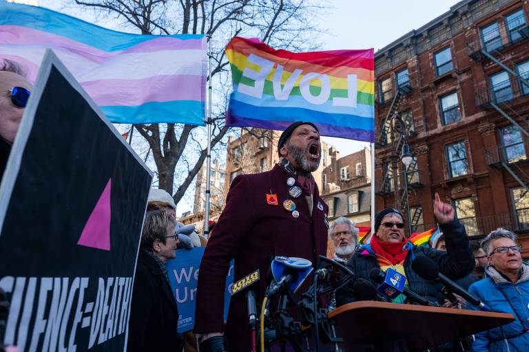 A imagem mostra uma manifestação com várias pessoas reunidas. Um homem em destaque， vestido com um casaco escuro e bottons coloridos， está falando em um microfone. Ao fundo， há bandeiras do orgulho LGBTQ+ e trans， além de cartazes com as palavras 039;SILENCE = DEATH039;. O ambiente é urbano， com prédios visíveis ao fundo.