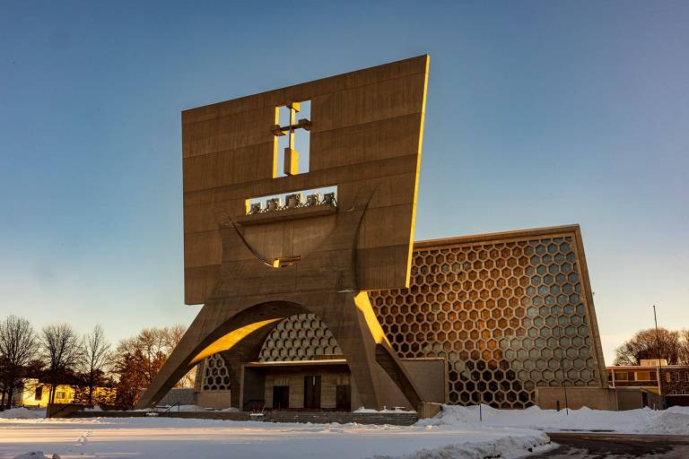 A imagem mostra uma igreja moderna com uma estrutura arquitetônica distinta. A fachada é composta por um grande painel retangular com uma cruz em destaque na parte superior. A base da igreja é ampla e possui formas geométricas， com um design que combina concreto e elementos hexagonais. O ambiente ao redor está coberto de neve， e o céu é claro， indicando um dia ensolarado.