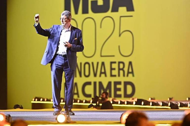 Um homem em um palanque apresenta um evento， com um fundo amarelo onde está escrito 039;MSA 2025 NOVA ERA CIMED039;. O homem está vestido com um terno escuro e levanta uma mão， enquanto a plateia é visível em primeiro plano， com várias pessoas assistindo.