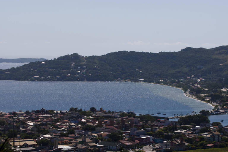 A imagem mostra uma vista panorâmica de uma baía cercada por colinas. No primeiro plano， há uma área urbana com várias casas e edifícios. A água da baía reflete a luz do sol， criando um efeito brilhante. Ao fundo， há uma ilha e colinas cobertas de vegetação.