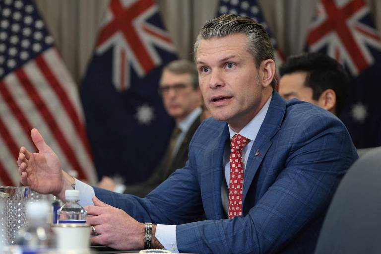 A imagem mostra um homem de terno azul com uma camisa branca e uma gravata vermelha， gesticulando enquanto fala em uma mesa de reunião. Ao fundo， há bandeiras da Austrália e outras pessoas visíveis， mas não identificáveis. O ambiente parece ser uma sala de conferências.