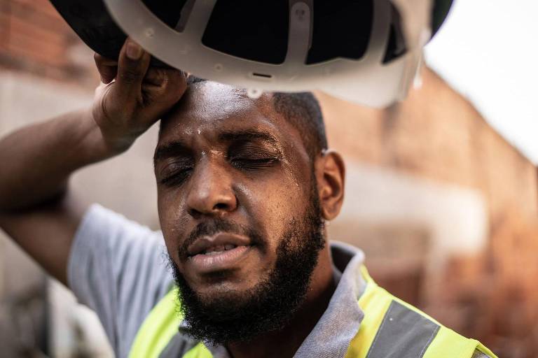 Um homem com pele escura e barba， vestindo uma camisa cinza e um colete amarelo， está segurando a cabeça com uma mão enquanto a outra mão segura um capacete. Ele parece estar suado e exausto， com os olhos fechados e uma expressão de cansaço. O fundo mostra uma parede de tijolos e um ambiente de construção.