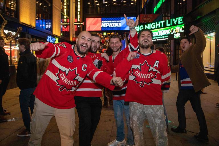 Um grupo de cinco homens está posando para a foto em uma área urbana iluminada à noite. Eles estão usando camisetas de hóquei vermelhas com o símbolo do Canadá. Os homens estão sorrindo e fazendo gestos de celebração， com alguns levantando os braços. Ao fundo， há letreiros luminosos e pessoas caminhando.