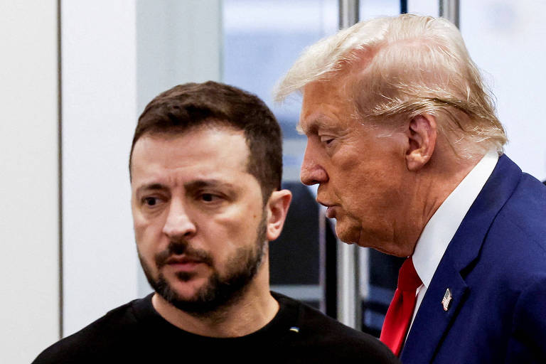 A imagem mostra dois homens em um ambiente interno. Um deles está de perfil， com cabelo escuro e barba， vestindo uma camiseta preta. O outro homem， com cabelo loiro e penteado， está inclinado em direção ao primeiro， parecendo falar com ele. O fundo é desfocado， mas sugere um ambiente de reunião.