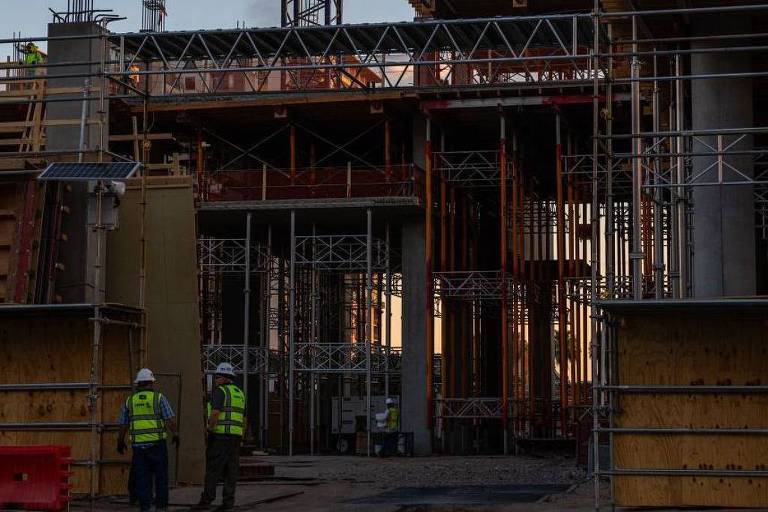 A imagem mostra um canteiro de obras com estruturas de andaimes e paredes em construção. Dois trabalhadores， vestidos com coletes de segurança amarelos， estão em pé em frente à entrada do edifício em construção. A iluminação é suave， sugerindo que a foto foi tirada ao entardecer.