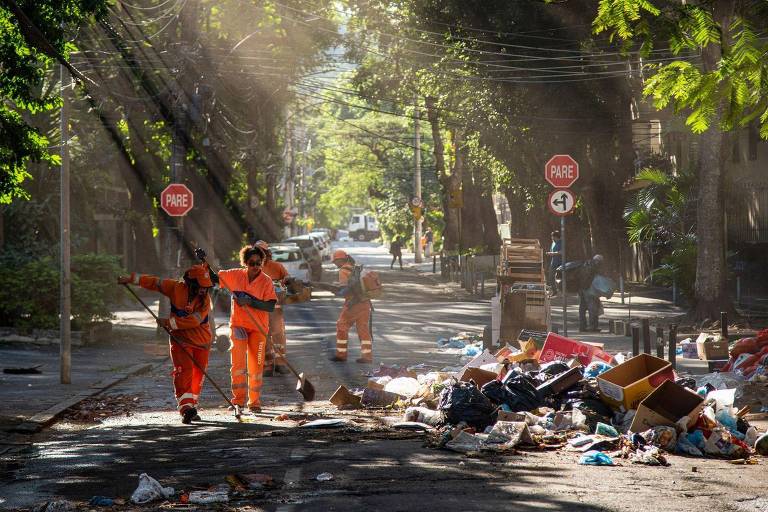 A imagem mostra uma rua arborizada com luz solar filtrando através das árvores. Dois trabalhadores em uniformes laranja estão limpando a rua， enquanto há lixo acumulado em um dos lados. Sinais de trânsito são visíveis ao fundo， e a cena transmite um ambiente de trabalho em equipe para a limpeza urbana.