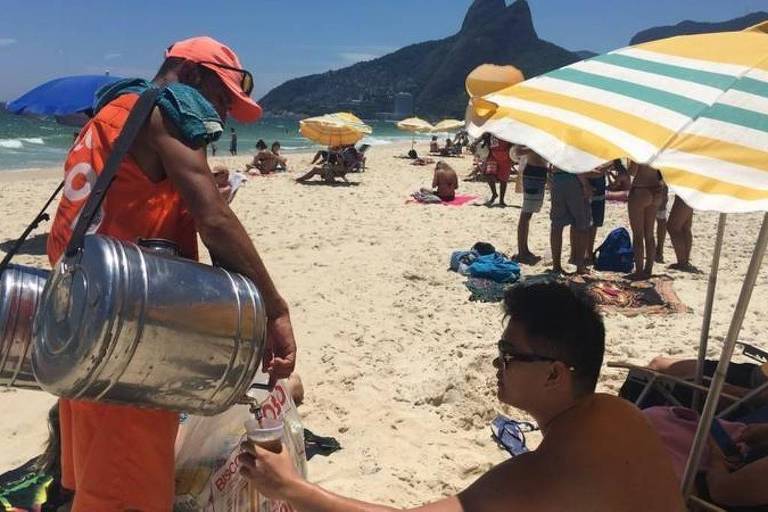 A imagem mostra um vendedor de bebidas na praia， usando um uniforme laranja e um chapéu. Ele está segurando um recipiente metálico grande， provavelmente com bebidas， enquanto se inclina em direção a um cliente sentado na areia. O cliente， que está usando óculos escuros， parece estar fazendo um pedido. Ao fundo， há várias pessoas relaxando sob guarda-sóis listrados e uma montanha ao longe.