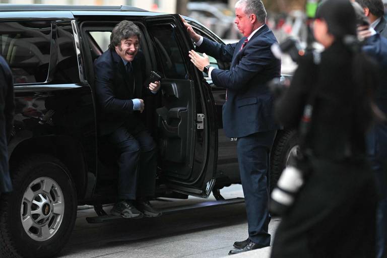 Um homem está saindo de um veículo preto， enquanto um segurança o ajuda a descer. O homem está vestido com um terno escuro e parece sorrir. O segurança está usando um terno claro e gesticula para ajudar o homem. Há pessoas ao fundo， algumas com câmeras.