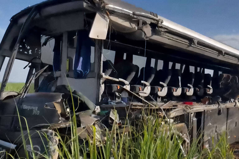 A imagem mostra um ônibus danificado， com a estrutura comprometida e parte da carroceria destruída. O ônibus está localizado em uma área com vegetação alta， possivelmente em um campo. O céu está claro， com algumas nuvens visíveis ao fundo.