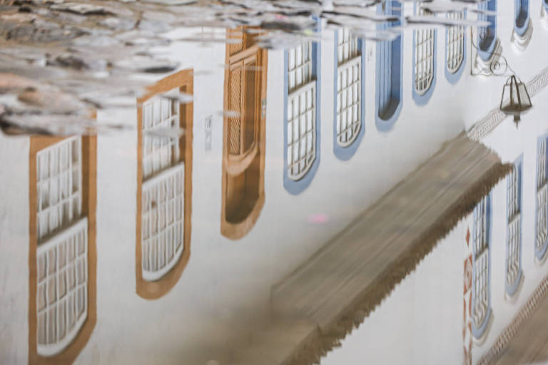 A imagem mostra o reflexo de um edifício em uma superfície de água. As janelas do edifício são brancas com molduras em tom de laranja e azul. O teto do edifício é coberto por uma estrutura que parece ser uma marquise. O reflexo é distorcido pela superfície da água.