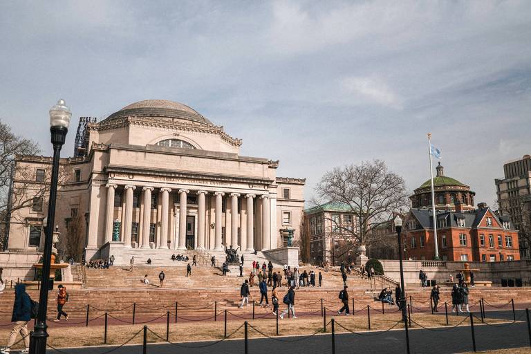 A imagem mostra a fachada da Universidade de Columbia， com um grande edifício de colunas e um domo. Há várias pessoas caminhando em frente ao edifício， que está situado em um espaço aberto com escadas. Ao fundo， é possível ver outro edifício com uma cúpula verde e árvores sem folhas， indicando a estação do ano. O céu está parcialmente nublado.