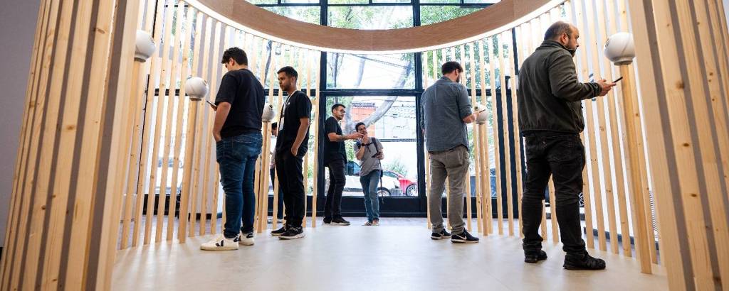 A imagem mostra um espaço de exposição com uma estrutura de madeira em forma de arco. Várias pessoas estão interagindo com a instalação, algumas observando e outras usando dispositivos móveis. Ao fundo, há grandes janelas que permitem a entrada de luz natural.