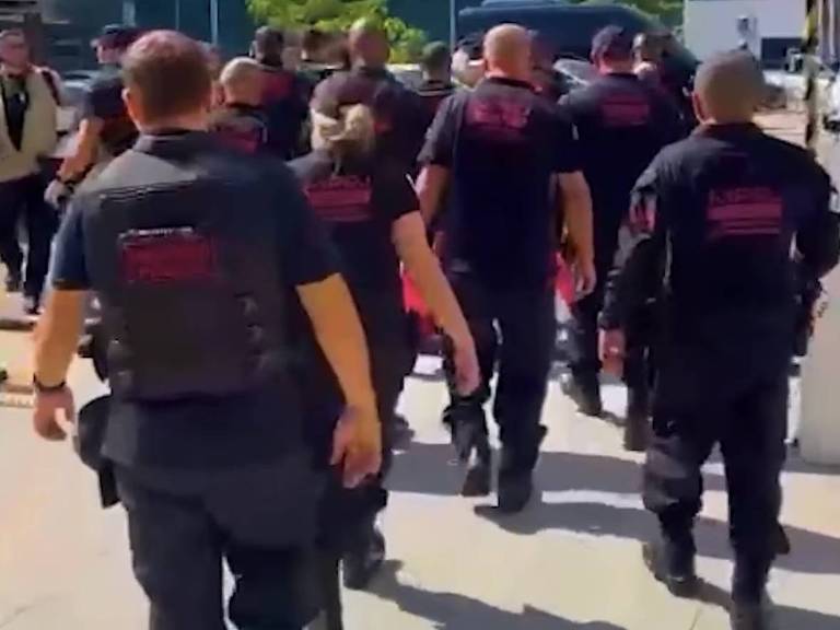 A imagem mostra um grupo de agentes de segurança ou policiais caminhando em direção a um local. Eles estão vestidos com uniformes pretos e coletes， e a maioria tem as costas voltadas para a câmera. O ambiente parece ser urbano， com edifícios ao fundo.