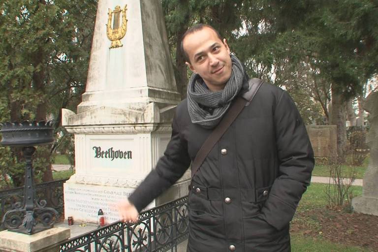A imagem mostra um homem em um cemitério, posando ao lado do túmulo de Beethoven. O túmulo é uma estrutura alta e branca, com o nome 'Beethoven' escrito em verde. Ao lado do túmulo, há um suporte de metal para velas. O homem está usando um casaco escuro e um cachecol, e parece sorrir enquanto aponta para o túmulo.