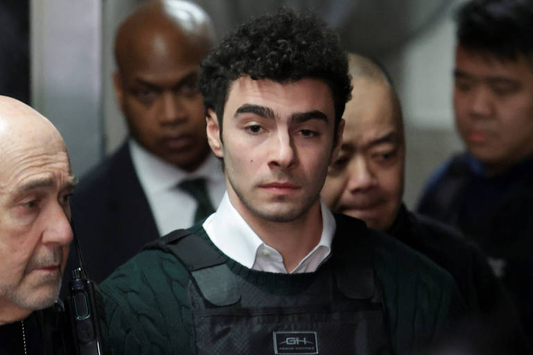 Um jovem com cabelo cacheado e escuro, vestindo um colete à prova de balas, está sendo acompanhado por homens em trajes formais. O ambiente parece ser um corredor de tribunal ou uma instalação de segurança. O jovem tem uma expressão séria e está olhando para frente, enquanto os outros homens ao seu redor observam atentamente.