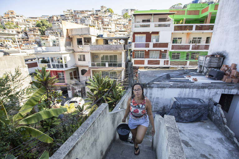 A imagem mostra uma mulher subindo uma escada em uma área urbana. Ela carrega um balde e está vestida com uma blusa colorida e shorts. Ao fundo， há várias casas empilhadas， com varandas e janelas， em um ambiente que parece ser uma comunidade. A vegetação， como plantas grandes， está visível ao lado da escada.