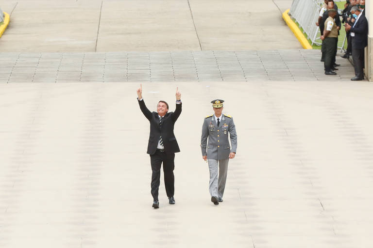 Um homem em um terno preto, com uma gravata listrada, está caminhando e levantando os braços em sinal de celebração. Ao seu lado, um oficial militar vestido com um uniforme cinza e um boné. O cenário é uma área pavimentada, com algumas pessoas ao fundo observando.