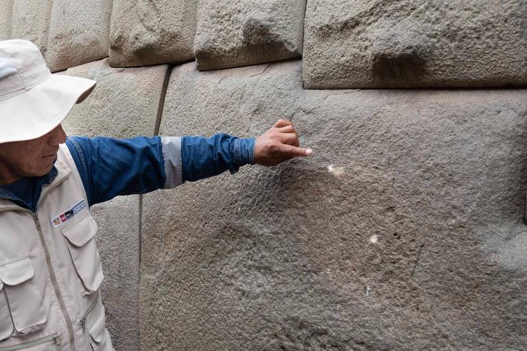 Um homem， vestindo um chapéu claro e uma camisa azul com detalhes brancos， está apontando para uma parede de pedra. A parede é composta por grandes blocos de pedra cinza， com algumas marcas visíveis na superfície. O homem parece estar explicando algo sobre a estrutura da parede.