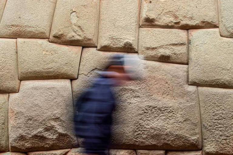 A imagem mostra uma parede composta por grandes blocos de pedra， com uma textura rugosa e irregular. Um indivíduo em movimento， desfocado， passa pela frente da parede， vestindo uma jaqueta escura e um boné. A parede parece antiga e é feita de pedras grandes e bem ajustadas.