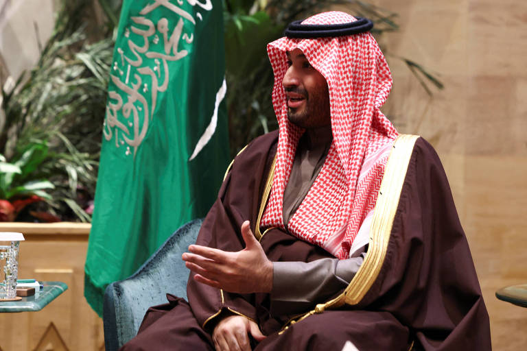 Um homem vestido com trajes tradicionais árabes， incluindo uma thobe e um ghutrah， está sentado em uma cadeira. Ele está gesticulando com a mão direita enquanto conversa. Ao fundo， há uma bandeira da Arábia Saudita e plantas decorativas.