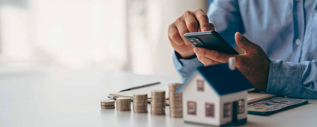 A imagem mostra uma pessoa segurando um smartphone com uma mão, enquanto a outra mão está apoiada na mesa. Ao lado, há uma pilha de moedas e uma pequena maquete de uma casa. Ao fundo, é possível ver um caderno e um ambiente iluminado.