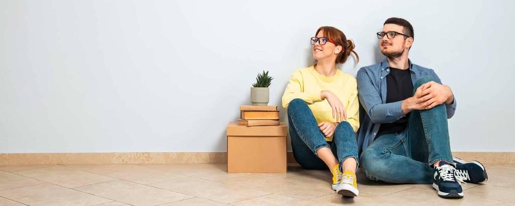 Um casal está sentado no chão de um ambiente interno com paredes claras. A mulher, com cabelo ruivo e óculos, usa uma blusa amarela e jeans. O homem, com cabelo escuro e óculos, veste uma camisa azul e jeans. Eles estão encostados na parede, olhando para direções opostas. Ao lado deles, há uma pequena planta em um vaso e algumas caixas empilhadas.