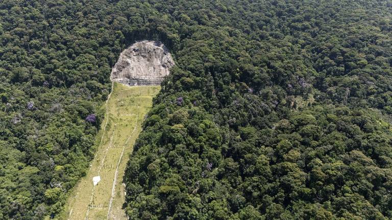 A imagem mostra uma área de floresta densa, com uma grande parte desmatada que revela uma superfície rochosa. A vegetação ao redor é verde e variada, enquanto a área desmatada apresenta um solo exposto e uma linha clara que se estende pela floresta.