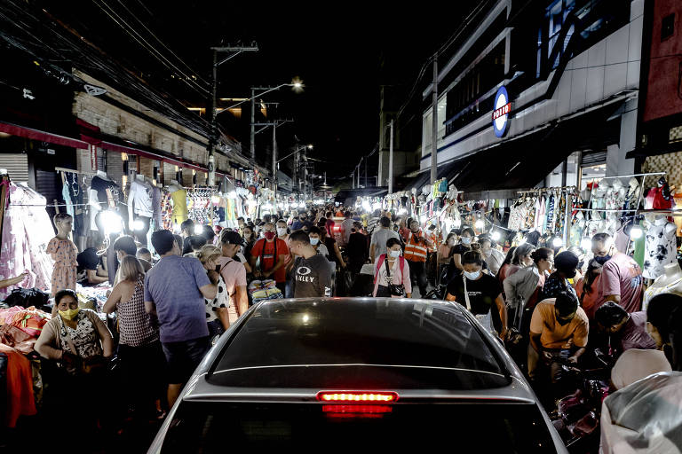 A imagem mostra um mercado noturno movimentado， com muitas pessoas circulando entre barracas iluminadas. Há uma grande quantidade de produtos expostos， e um carro está estacionado na frente， dificultando a passagem. As luzes das barracas criam um ambiente vibrante e animado.
