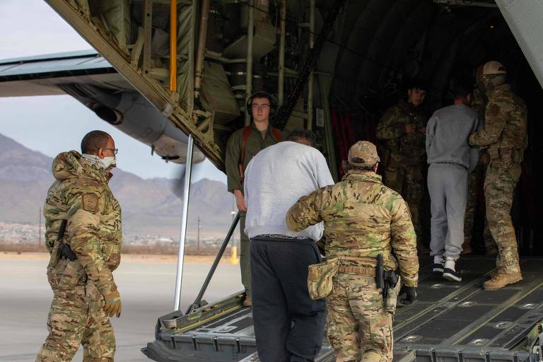 A imagem mostra um grupo de militares em um avião de transporte. Alguns soldados estão de pé na entrada do avião， enquanto outros estão dentro. Um dos soldados está ajudando uma pessoa vestindo roupas escuras a entrar no avião. O ambiente é ao ar livre， com montanhas ao fundo e um céu claro.