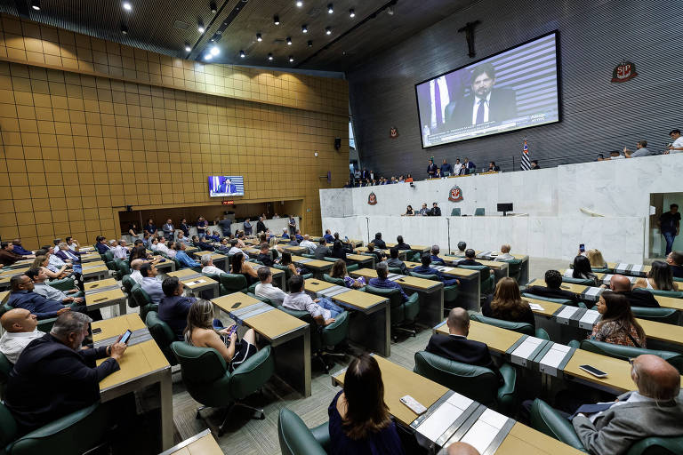 A imagem mostra uma sessão legislativa em um ambiente formal. Há várias pessoas sentadas em mesas dispostas em fileiras， com algumas assistindo a uma apresentação em um telão. No fundo， um grupo de pessoas está em pé， observando. O ambiente é bem iluminado， com paredes de cor clara e um design moderno. A mesa principal está ocupada por alguns indivíduos， e há um símbolo visível na parede.