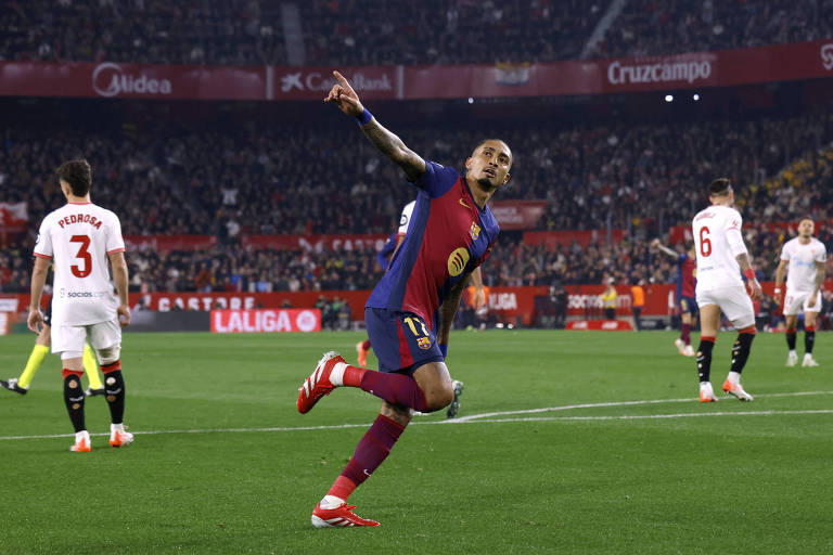 Um jogador do Barcelona， vestindo um uniforme vermelho e azul， celebra após marcar um gol. Ele está em campo， com um gesto de comemoração， enquanto outros jogadores do time adversário estão ao fundo. O estádio está cheio de torcedores， e há banners visíveis nas arquibancadas.