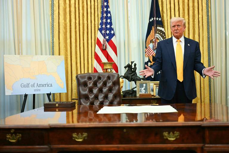 A imagem mostra um homem em pé ao lado de uma mesa de escritório， com uma bandeira dos Estados Unidos ao fundo. Ele está usando um terno escuro e uma gravata amarela. À esquerda， há um quadro com um mapa da região do Golfo da América， e na mesa há um documento em branco. O ambiente é decorado com cortinas douradas e um brasão presidencial.