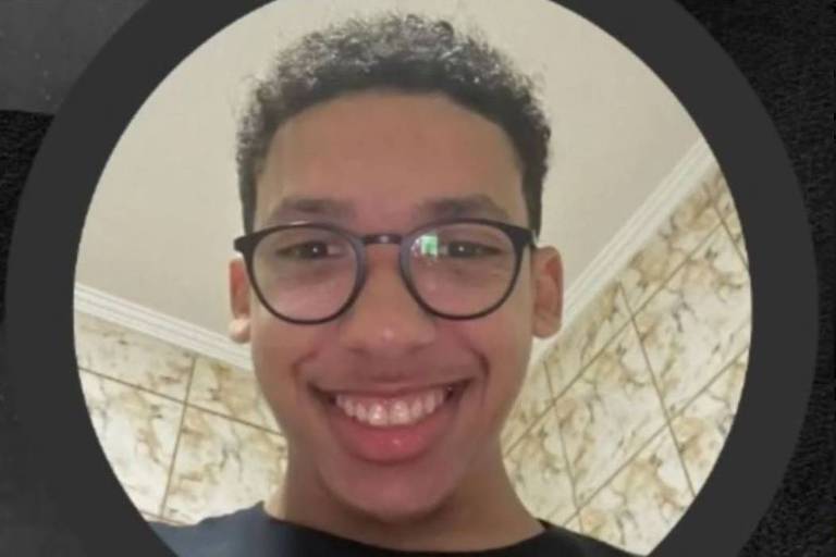 A imagem mostra um jovem com cabelo cacheado e óculos， sorrindo para a câmera. O fundo é uma parede com azulejos claros e um padrão discreto. A foto é circular e destaca o rosto do jovem.