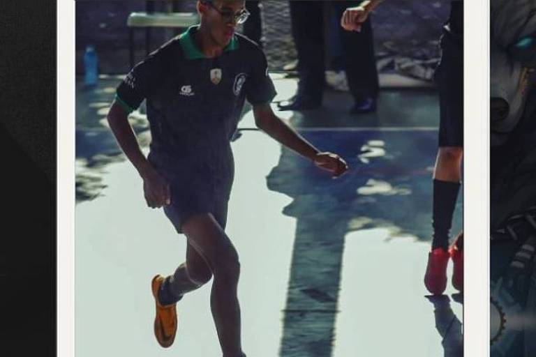 A imagem mostra um jogador de futebol em movimento， vestindo um uniforme escuro e chuteiras laranjas. Ele está correndo em direção à câmera， com uma expressão focada. Ao fundo， há pessoas e uma superfície que parece ser um campo ou quadra， refletindo luz.