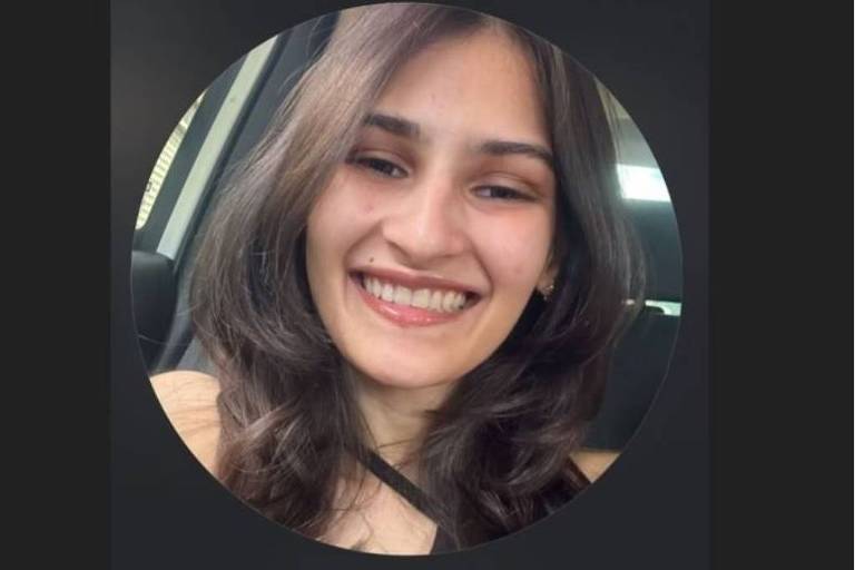 A imagem mostra o rosto de uma jovem mulher sorrindo. Ela tem cabelos longos e ondulados， e está usando uma blusa de alças. O fundo é desfocado， sugerindo que ela está dentro de um carro.