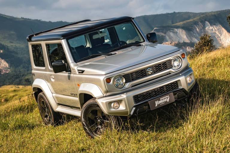 A imagem mostra um veículo SUV Suzuki Jimny prateado estacionado em um campo verde， com montanhas ao fundo. O carro tem um teto preto e está em uma posição inclinada， destacando suas rodas pretas e design robusto. O céu está parcialmente nublado， e a vegetação ao redor é densa.