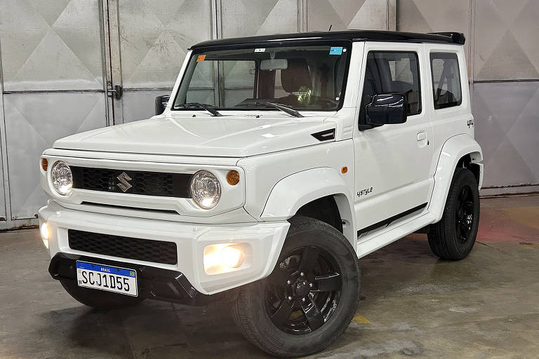 Imagem de um veículo Suzuki Jimny na cor branca, estacionado em um ambiente interno. O carro possui rodas pretas e detalhes em preto, com faróis dianteiros redondos e uma grade frontal distinta. O interior é visível através das janelas, mostrando um painel simples. O fundo da imagem é uma parede metálica com painéis de alumínio.