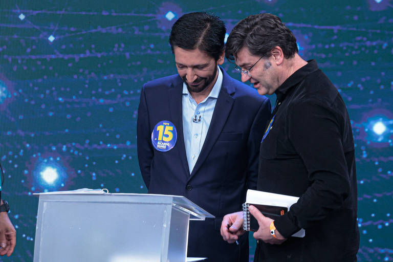 Dois homens estão em um estúdio de televisão， inclinando-se sobre uma mesa transparente. Um deles está usando um terno escuro e uma camisa clara， enquanto o outro veste uma camisa preta. Ambos parecem estar discutindo ou analisando algo. Ao fundo， há um cenário com luzes e efeitos visuais.