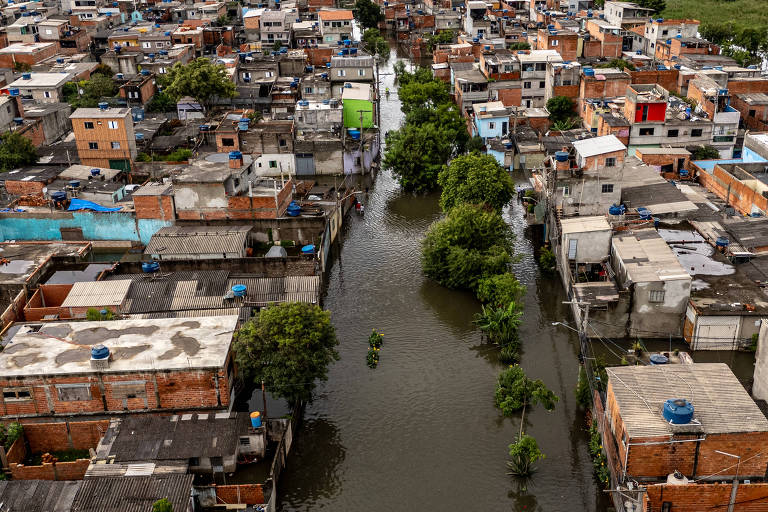 Imagem aérea de uma área urbana com casas e ruas alagadas. A água cobre as ruas， e algumas árvores estão visíveis， crescendo em meio à inundação. As casas são de diferentes cores， com telhados de diversos materiais. A cena mostra a gravidade da inundação em uma comunidade.