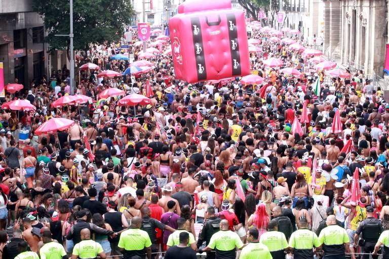 Uma grande multidão participa de uma parada de carnaval， com pessoas vestidas de forma colorida e festiva. Há um grande inflável rosa em forma de mala no centro da imagem， cercado por várias sombrinhas vermelhas. A cena é animada， com muitos foliões e alguns policiais na frente， observando a festa.