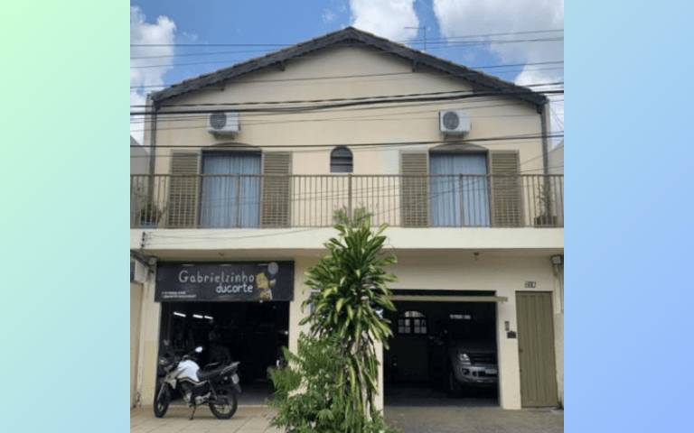 A imagem mostra a fachada de um edifício de dois andares. O prédio é de cor clara, com janelas com persianas verdes e uma varanda na parte superior. Na parte inferior, há uma entrada de garagem e uma loja com uma placa visível. À frente, há uma planta verde e uma bicicleta estacionada.