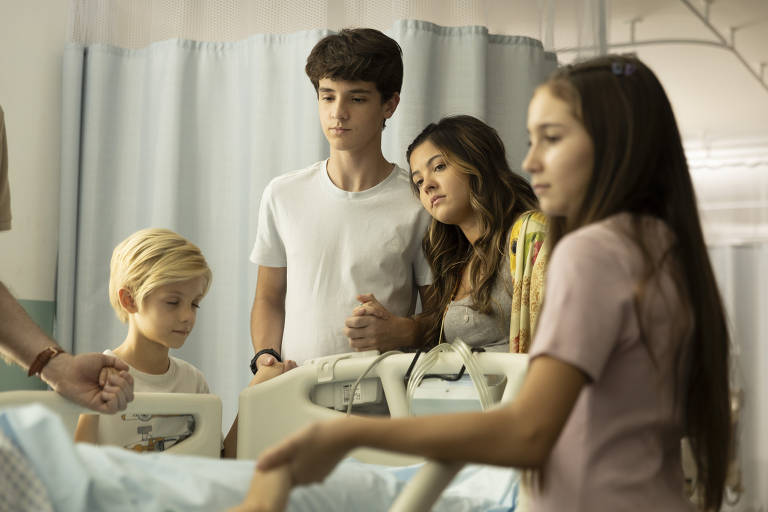 A imagem mostra um grupo de quatro jovens em um ambiente hospitalar. Eles estão em frente a uma cama de hospital, onde uma pessoa está deitada, mas não é visível. O grupo é composto por um menino loiro, um adolescente com cabelo castanho, uma jovem com cabelo castanho e uma menina com cabelo longo. Todos parecem preocupados e atentos à situação. O ambiente é iluminado e há cortinas azuis ao fundo.