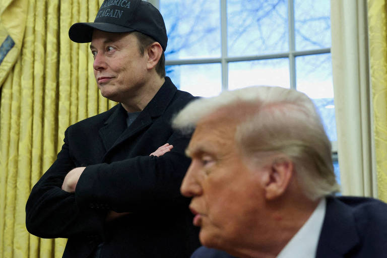 A imagem mostra um homem com um boné preto e uma jaqueta escura， com os braços cruzados， observando um outro homem que está de perfil， com cabelo loiro e usando um terno claro. O fundo é composto por cortinas amarelas e janelas， sugerindo um ambiente interno formal.