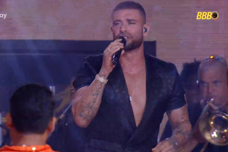 Um cantor está se apresentando em um palco, segurando um microfone. Ele usa uma camisa preta com um decote profundo e tem tatuagens visíveis nos braços. Ao fundo, há músicos e instrumentos, e a iluminação é vibrante. O ambiente parece ser de um evento ao vivo.