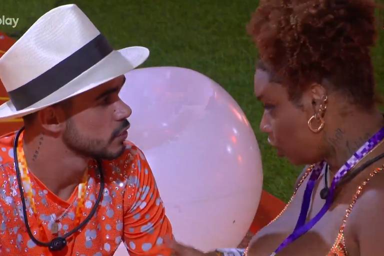 A imagem mostra dois participantes em um ambiente descontraído. Um homem com um chapéu branco e uma camisa laranja com estampas está olhando para uma mulher com cabelo cacheado e uma blusa brilhante. Eles parecem estar em uma conversa íntima, com um grande balão branco ao fundo.